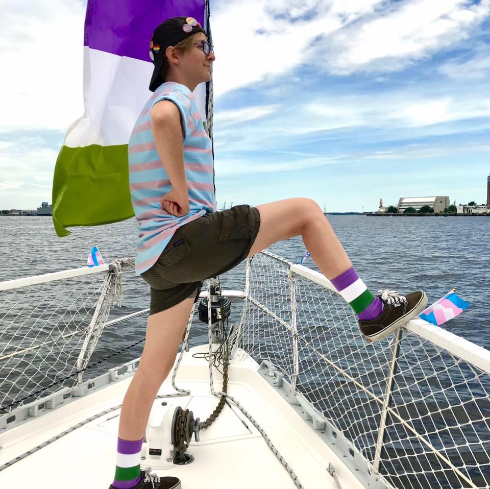Hunter Noffsinger standing in front of the ocean on a boat looking out with her gender queer flag socks on and a very happy expression